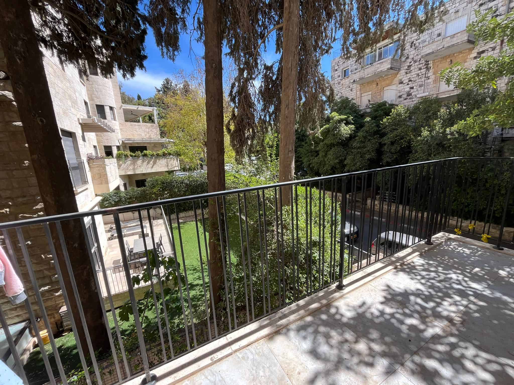 Balcony railing with shadows overlooking a courtyard, flanked by stone apartment buildings and tall trees.