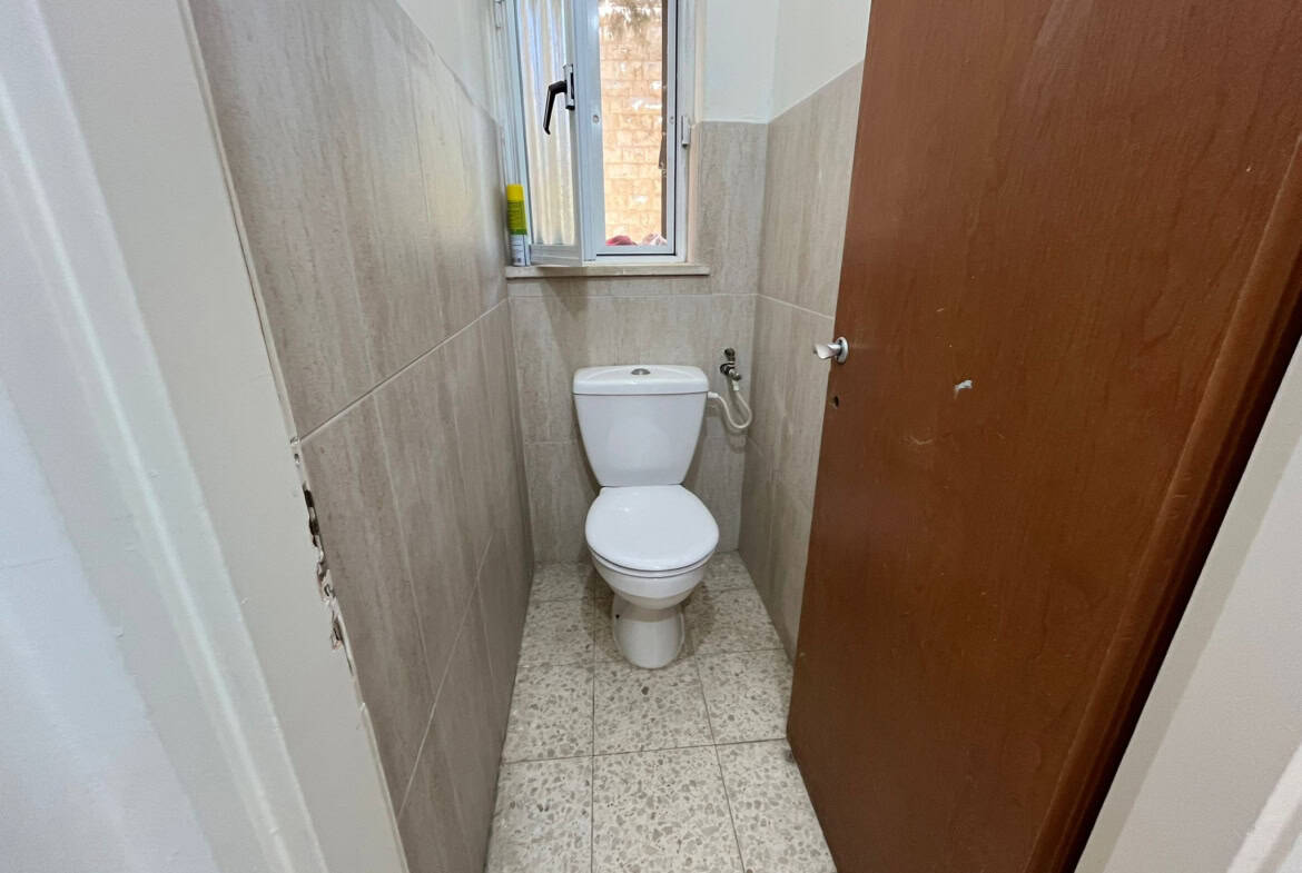 Narrow bathroom with a white toilet, beige tiled walls, and a window above the tank; a yellow bottle sits on the window ledge; wooden door on the right is open.