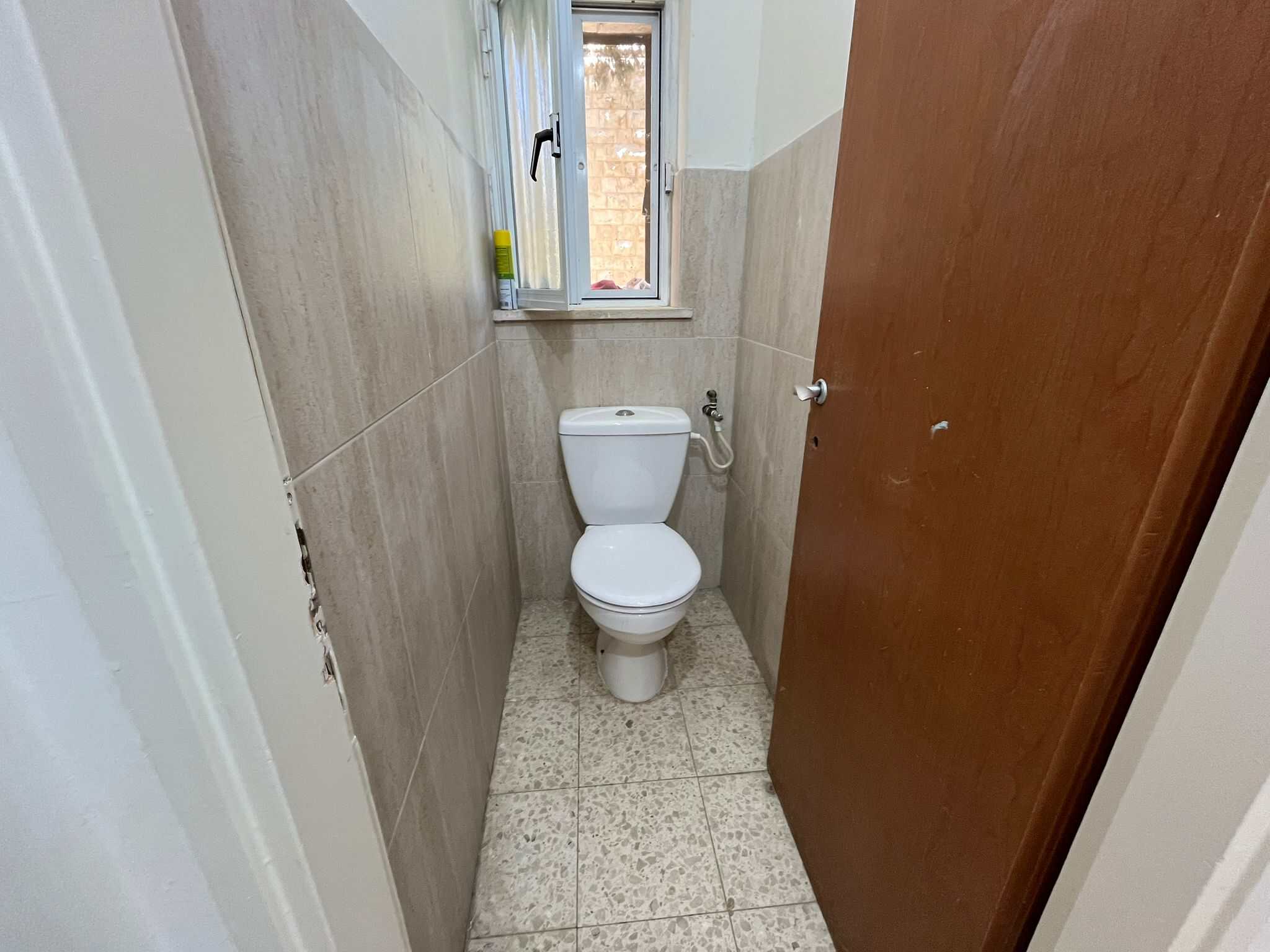 Narrow bathroom with a white toilet, beige tiled walls, and a window above the tank; a yellow bottle sits on the window ledge; wooden door on the right is open.