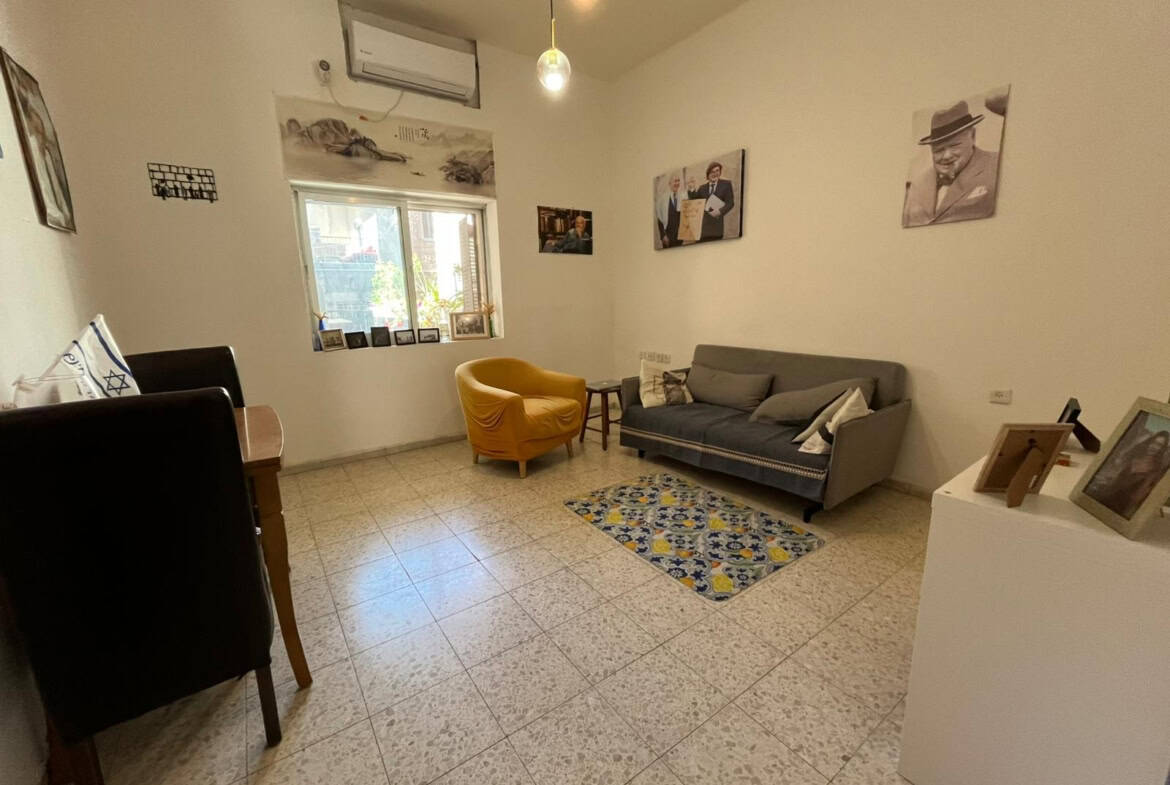 Cozy living room with a gray sofa, a mustard-yellow armchair, a small table, and a patterned rug; window with plants outside and wall art above.