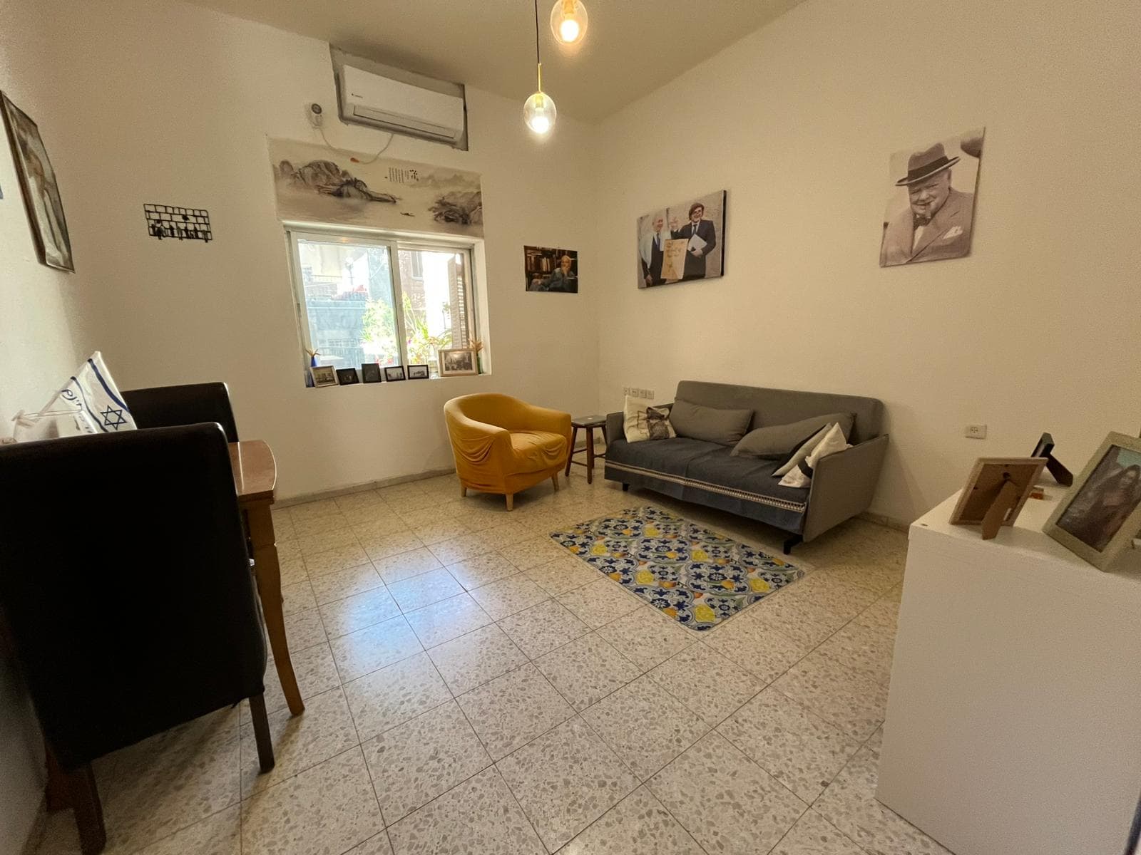 Cozy living room with a gray sofa, a mustard-yellow armchair, a small table, and a patterned rug; window with plants outside and wall art above.