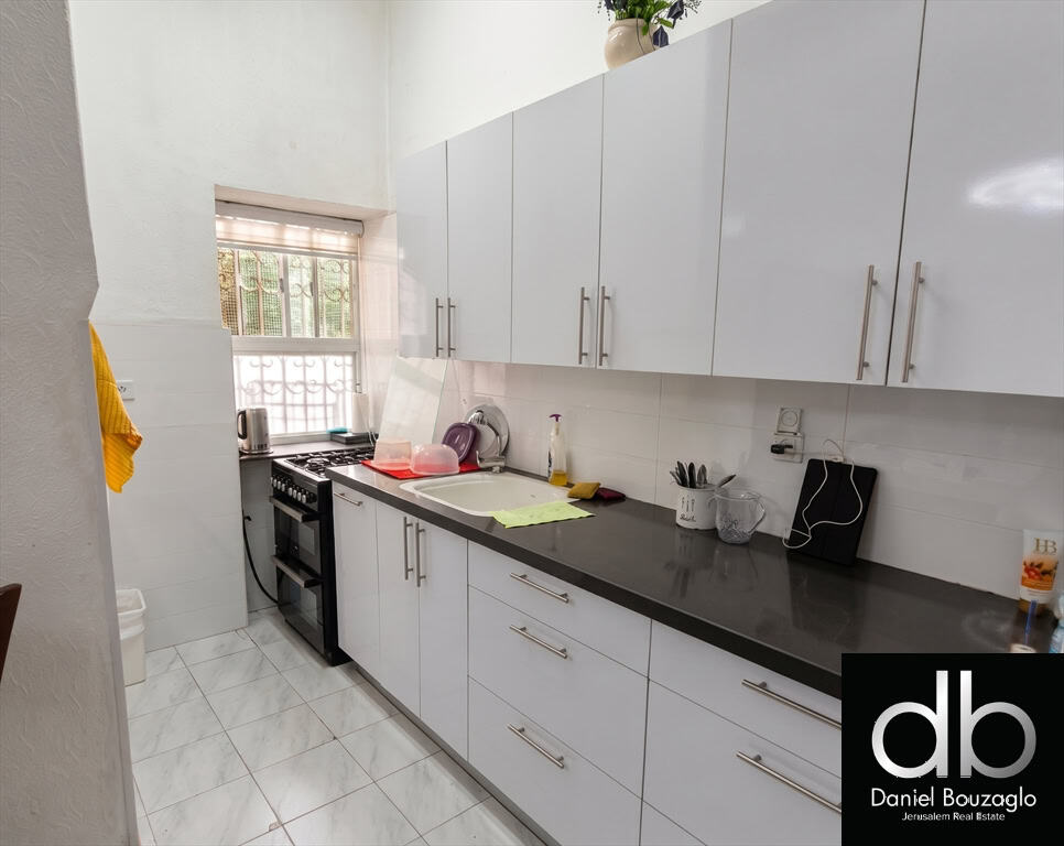 Long narrow kitchen with white glossy cabinets, dark countertop, sink, dish rack, and stove by a window.