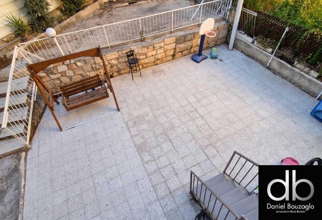 Spacious tiled backyard patio with a wooden porch swing, a small stool, a basketball hoop, and a stone retaining wall; watermark in bottom-right corner.