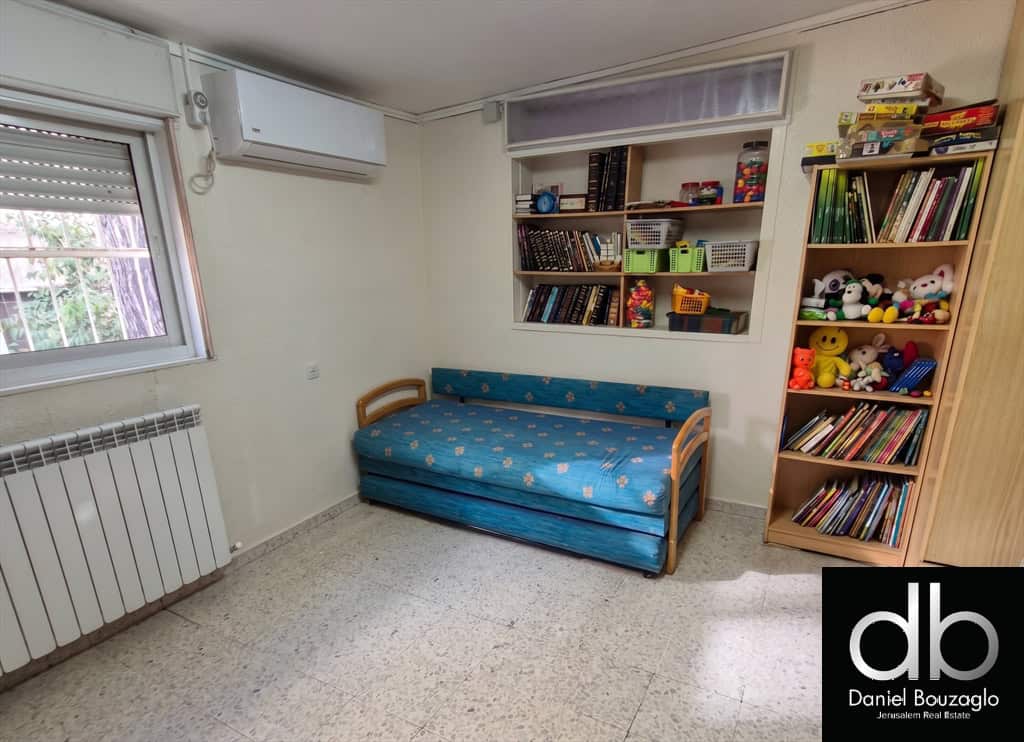 Small children's room with a blue fold-out sofa bed, white walls, and a window with a radiator below; shelves hold books and stuffed animals, plus a tall toy/book shelf on the right; branding watermark bottom right.