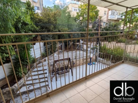 Balcony view overlooking a courtyard with stairs, a wooden swing bench, plants, and trees; real estate logo bottom right.
