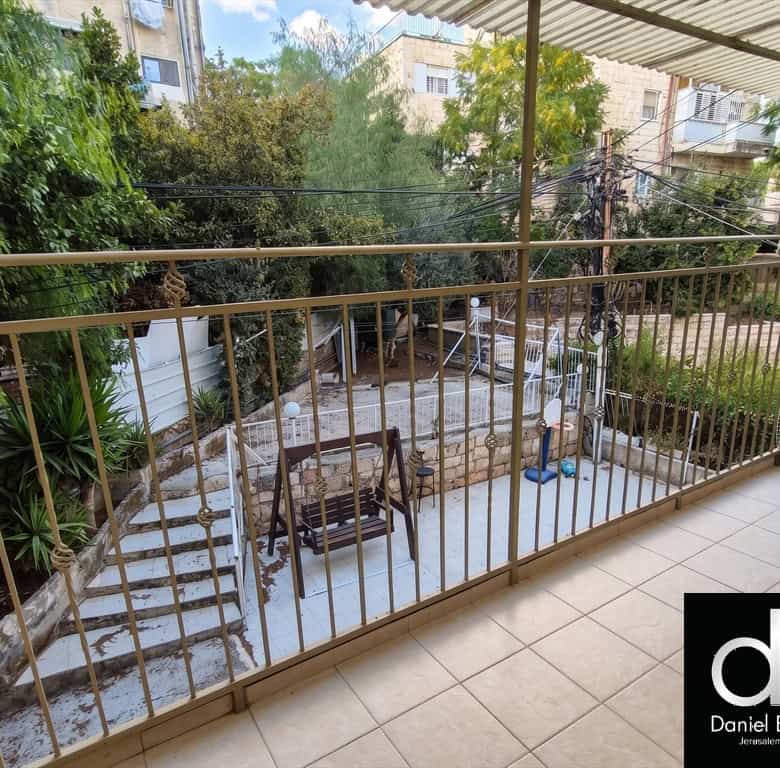 Balcony view overlooking a courtyard with stairs, a wooden swing bench, plants, and trees; real estate logo bottom right.