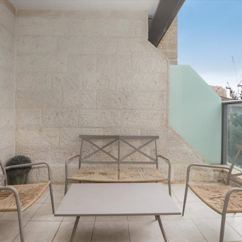 Balcony seating area with three metal-framed chairs with woven seats around a rectangular coffee table, beige stone wall, and glass railing with city buildings beyond.