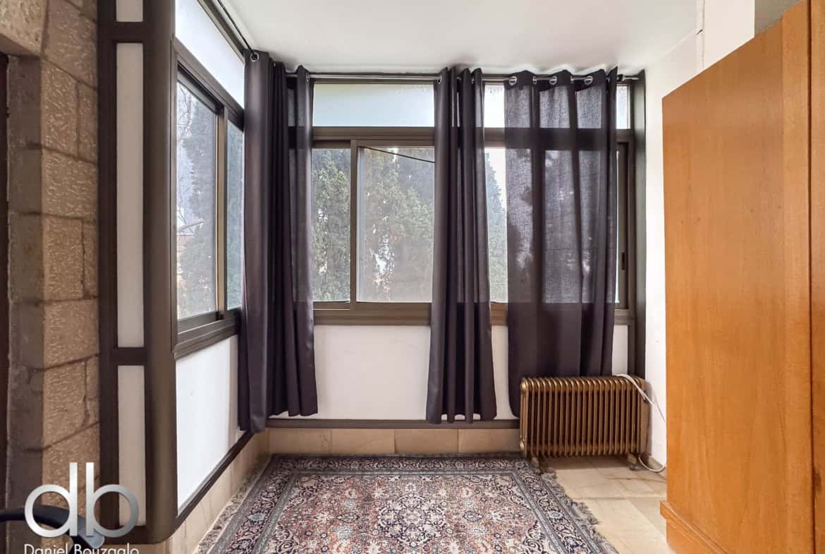 Interior hallway featuring a large window with dark curtains, a stone wall on the left, and a wooden panel on the right with a radiator beneath the window.