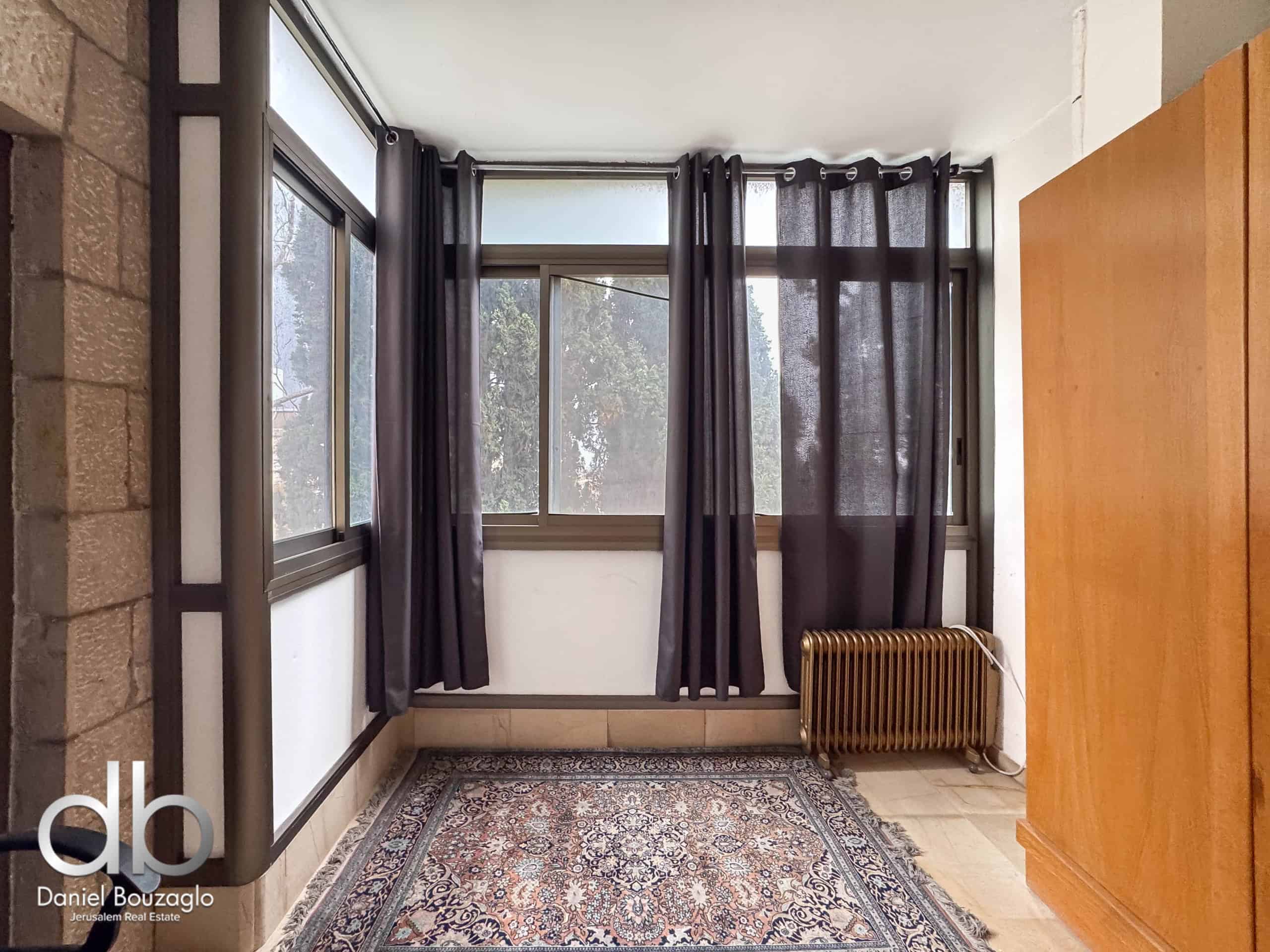 Interior hallway featuring a large window with dark curtains, a stone wall on the left, and a wooden panel on the right with a radiator beneath the window.