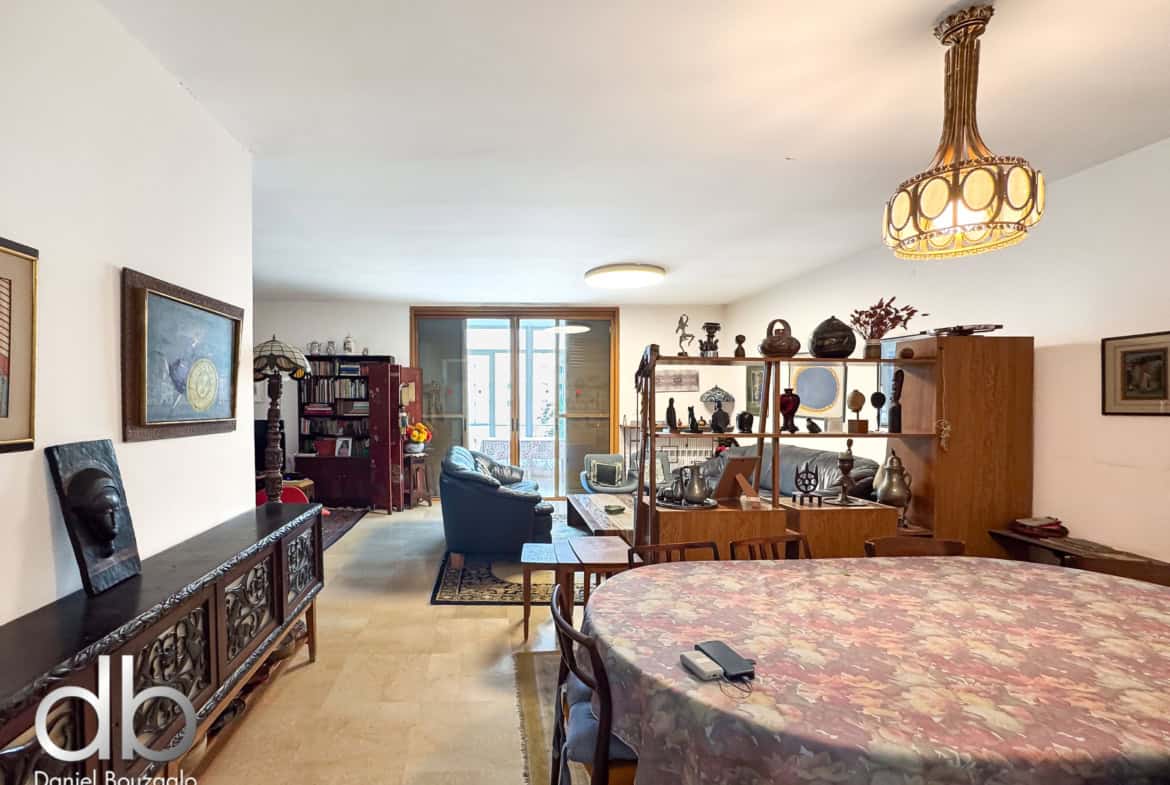 Living room filled with antique furniture and display shelves, a large patterned table in the foreground, and an ornate chandelier overhead.