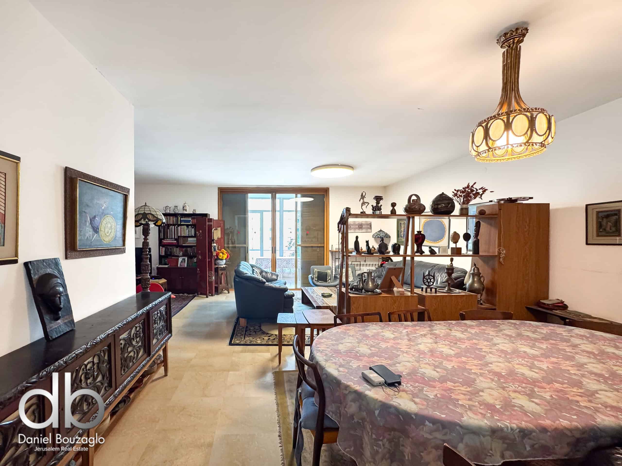 Living room filled with antique furniture and display shelves, a large patterned table in the foreground, and an ornate chandelier overhead.