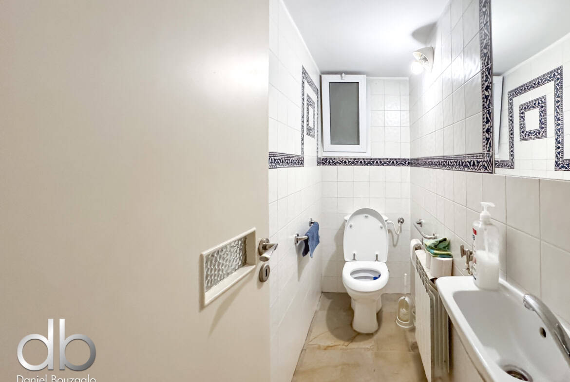 Narrow white-tiled bathroom with a toilet in the center, sink and radiator along the right wall, and blue decorative tile borders; small window above the toilet.