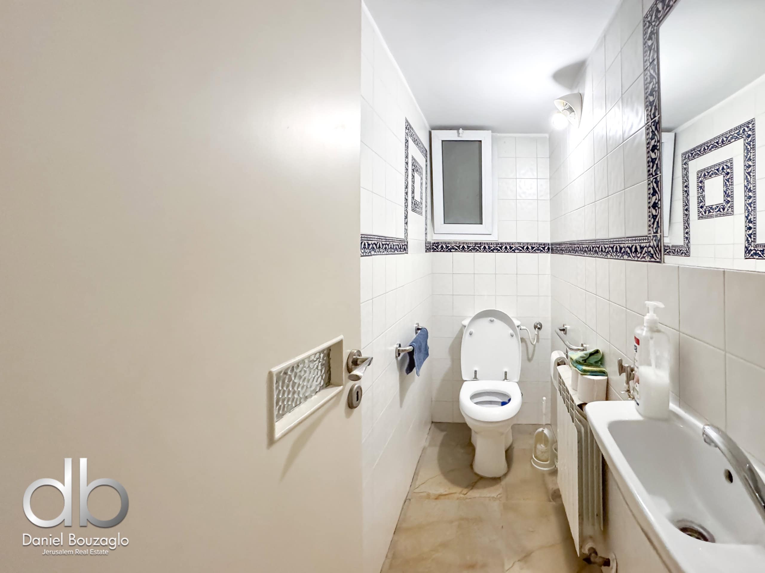 Narrow white-tiled bathroom with a toilet in the center, sink and radiator along the right wall, and blue decorative tile borders; small window above the toilet.