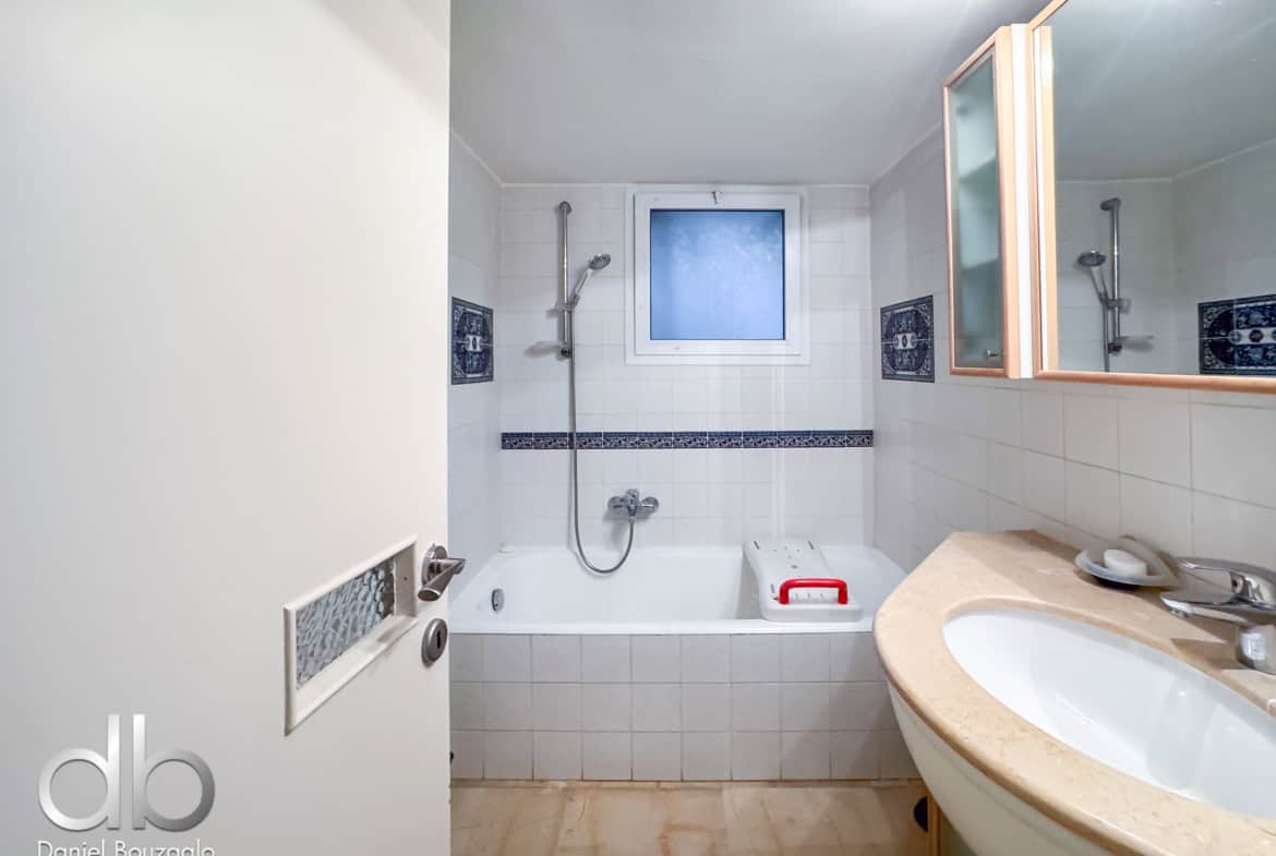 White tiled bathroom with a built-in bathtub, wall-mounted shower and handheld shower, frosted window above the tub, and a beige countertop with a sink and mirror cabinet.