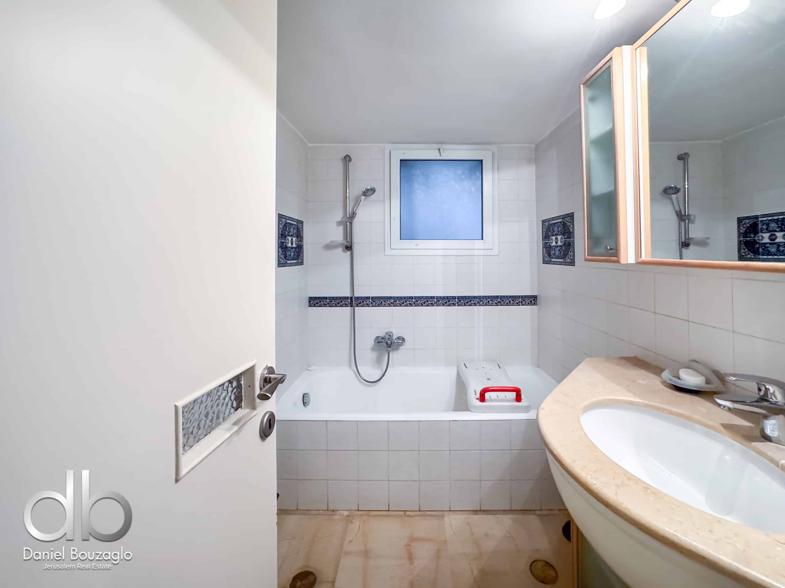 White tiled bathroom with a built-in bathtub, wall-mounted shower and handheld shower, frosted window above the tub, and a beige countertop with a sink and mirror cabinet.