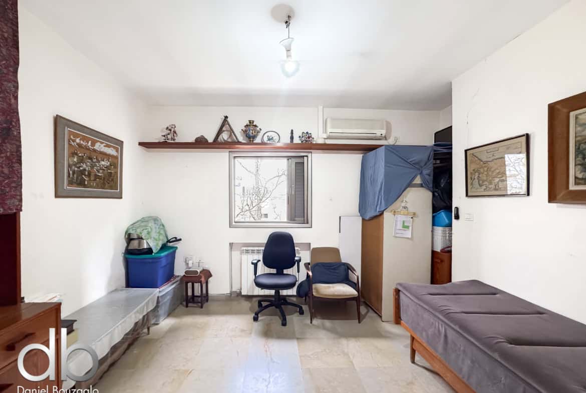 Living room with white walls, a wooden shelf of decorations above a window, and a blue office chair facing a desk area on the left.