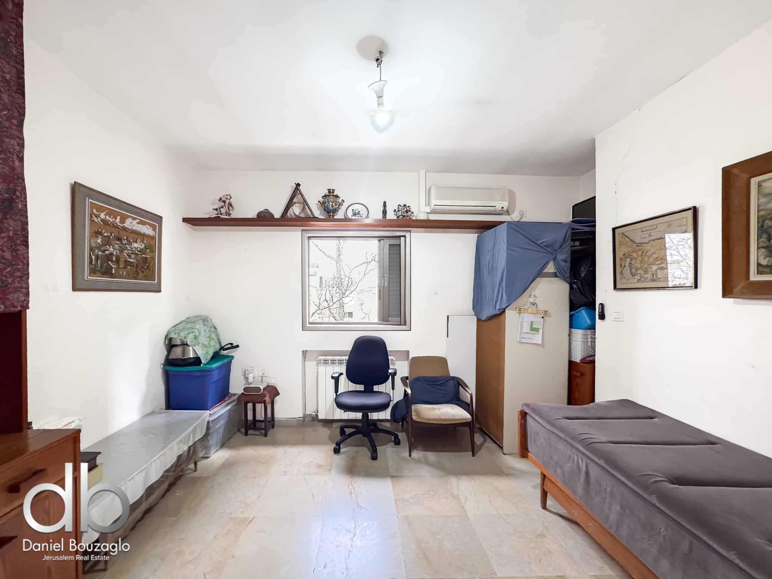 Living room with white walls, a wooden shelf of decorations above a window, and a blue office chair facing a desk area on the left.