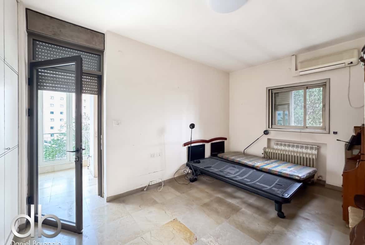 Sunlit living room with a treadmill along the right wall, glass balcony door on the left, and a window with an air conditioner above.
