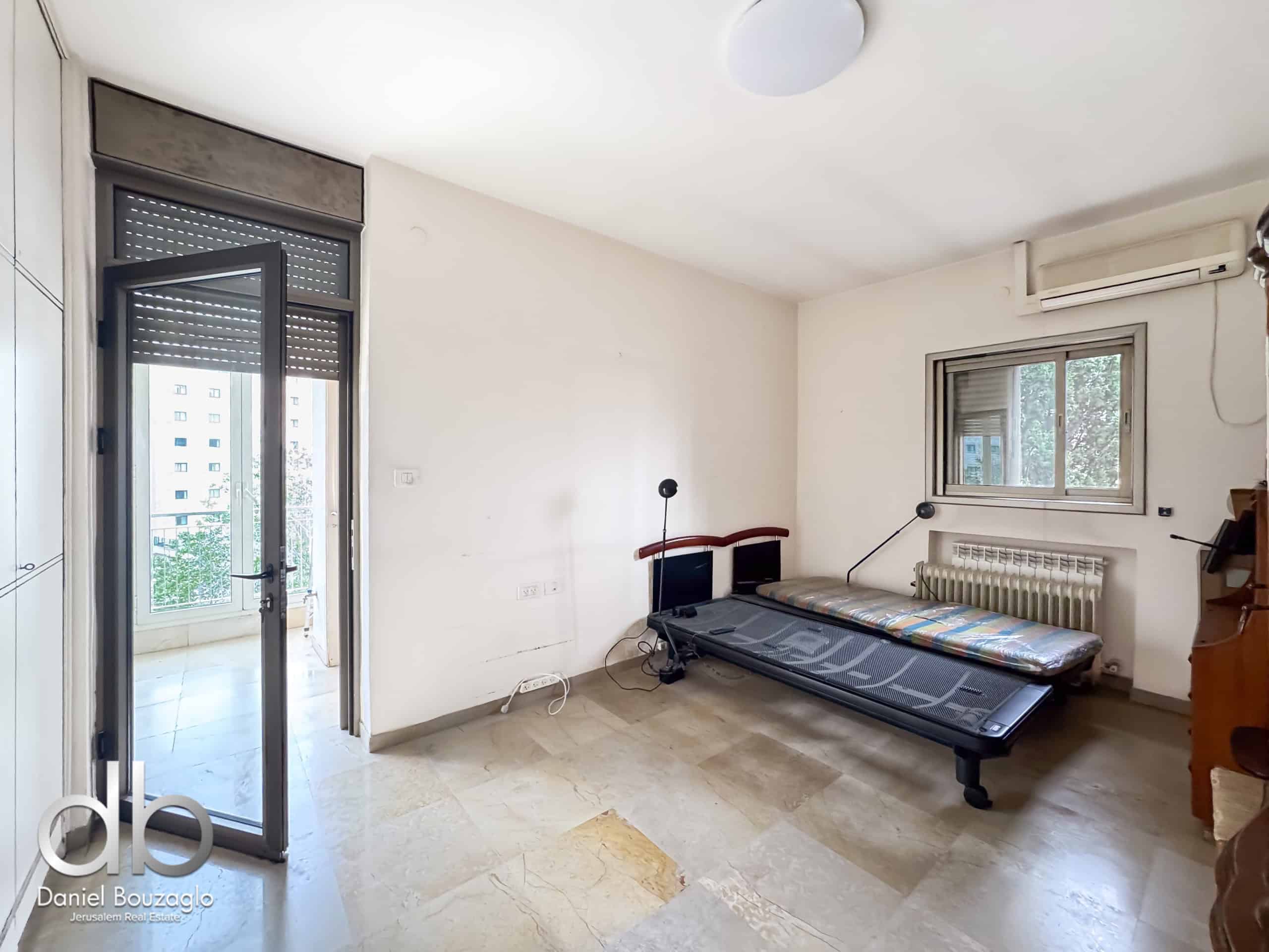 Sunlit living room with a treadmill along the right wall, glass balcony door on the left, and a window with an air conditioner above.