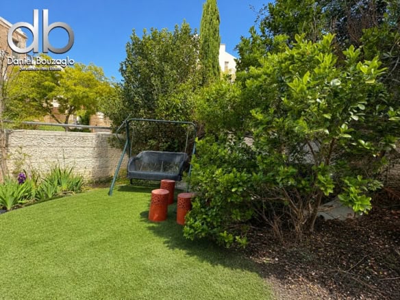 Backyard garden with a metal swing seat on a green lawn, three orange perforated stools, and dense shrubs beside a stone wall; real estate logo in top-left corner.