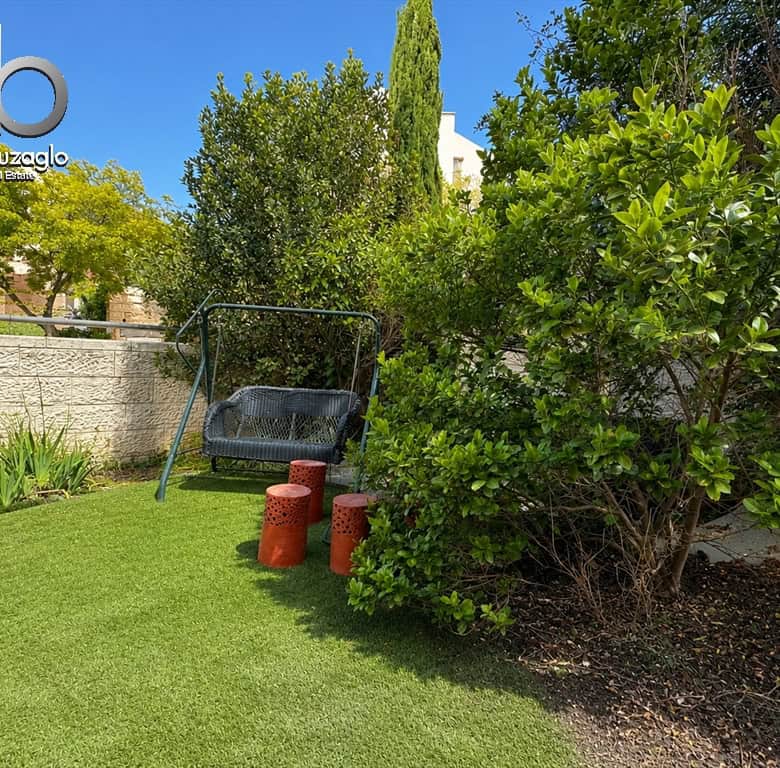 Backyard garden with a metal swing seat on a green lawn, three orange perforated stools, and dense shrubs beside a stone wall; real estate logo in top-left corner.