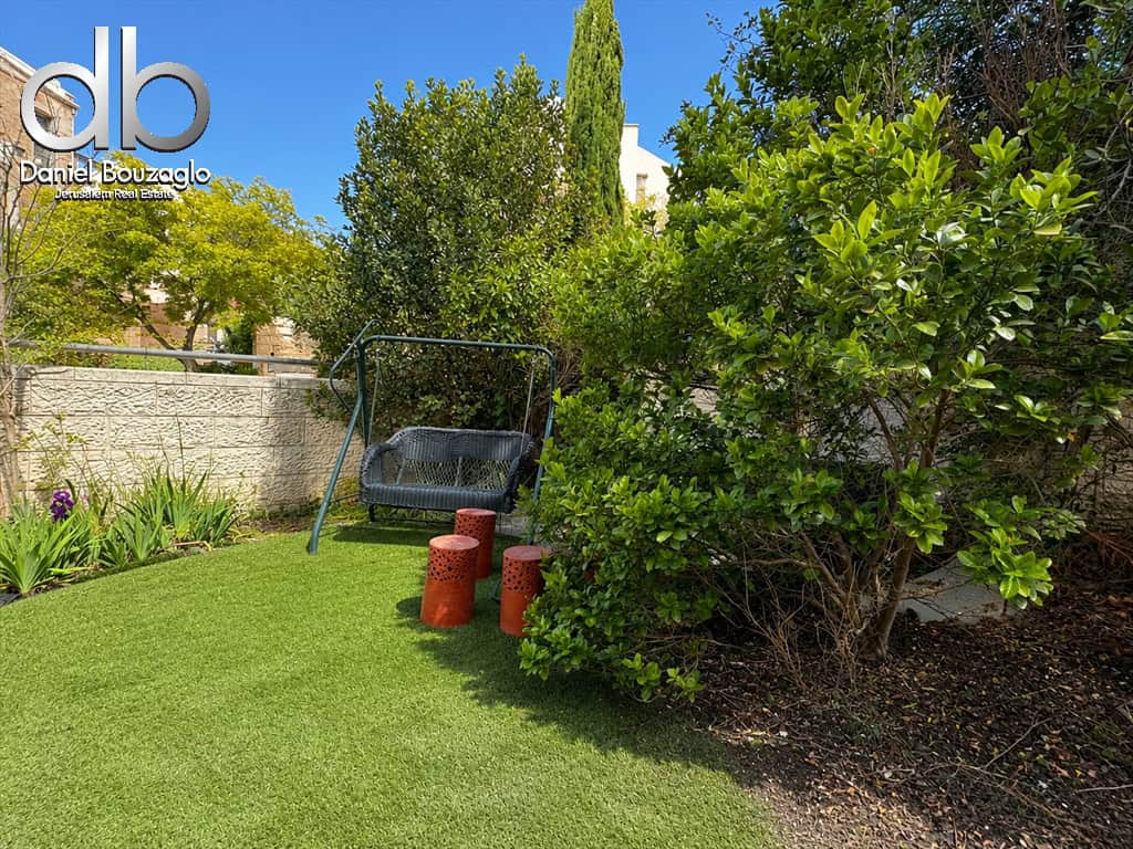 Backyard garden with a metal swing seat on a green lawn, three orange perforated stools, and dense shrubs beside a stone wall; real estate logo in top-left corner.