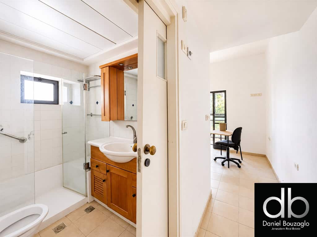 Bright bathroom with a glass shower, white tiles, and a wooden vanity; doorway opens to a small office area with a desk and chair in natural light.
