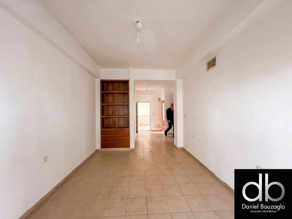 Empty living room with beige tile floor, white walls, and a built-in wooden shelf unit; a man stands near a doorway in the distance.