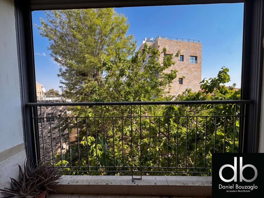 Balcony view with a metal railing, lush green tree, and a beige brick building against a clear blue sky.