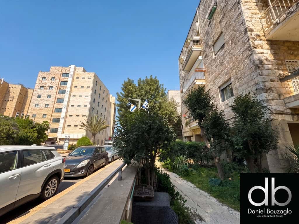 Urban street scene with mid-rise beige buildings, parked cars along the curb, and greenery under a clear blue sky.
