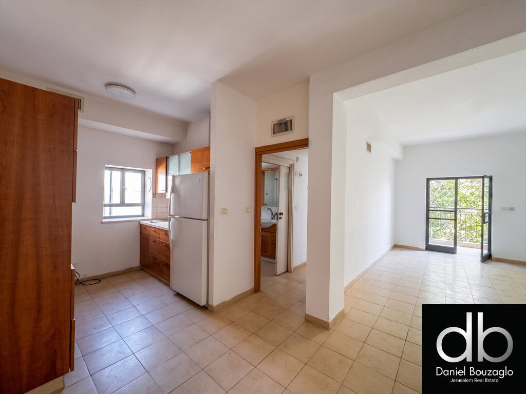 Bright kitchen area with wooden cabinets, white refrigerator, and a window overlooking outdoors; open to a tiled living space and doorway to a bathroom.