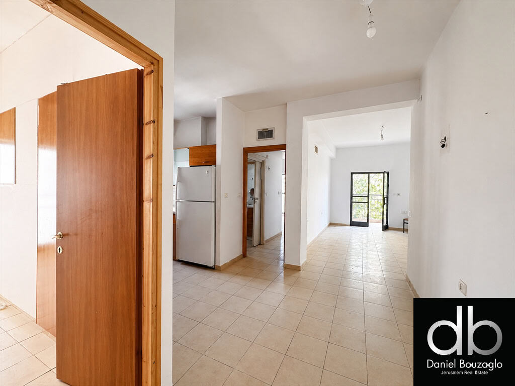 Empty open-plan apartment with tiled floor, white walls, and a balcony door at the far end; fridge visible in the kitchen area to the left.