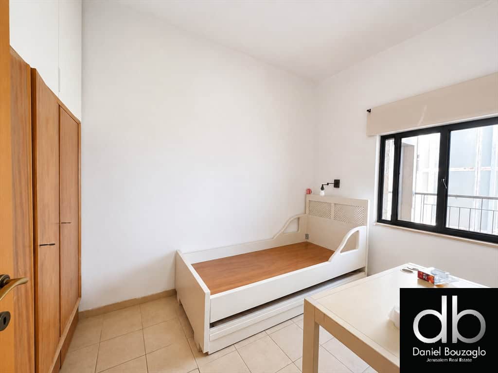 Small white bedroom with a single bed, a wooden wardrobe on the left, and a window to the right letting in light.