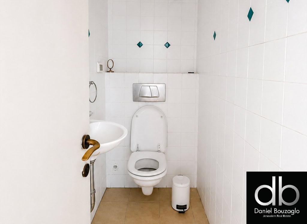 Compact bathroom with a wall-mounted toilet, small sink on the left, beige floor tiles, white tiled walls with teal diamond accents, and a logo corner.