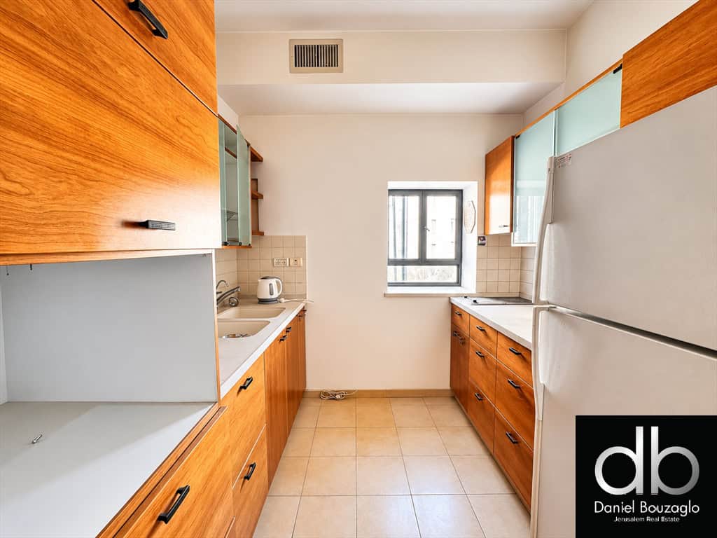 Narrow kitchen with wooden cabinets, white countertops, and a refrigerator on the right; window at the far wall adds natural light.