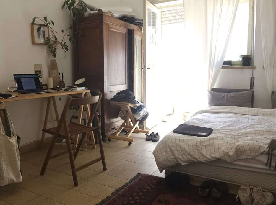 A tidy-looking bedroom with a wooden desk, laptop, and chair on the left, a large wardrobe, and an open door letting bright light into the room.