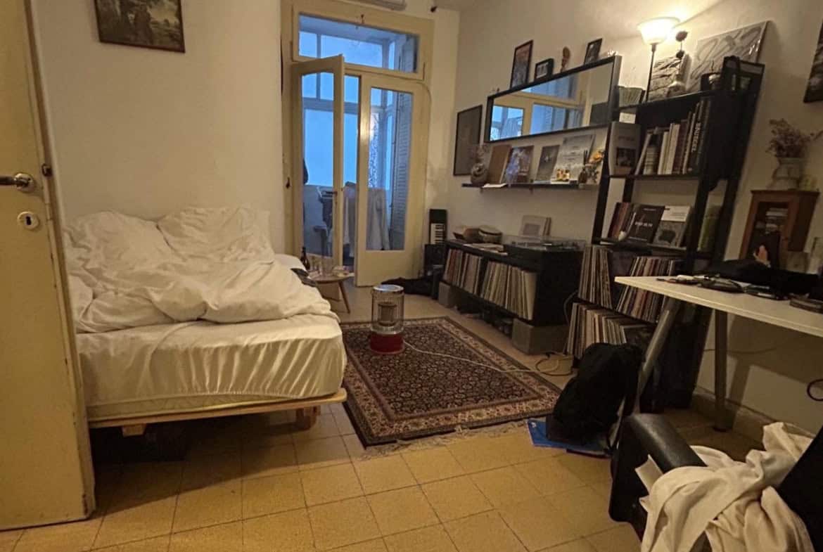 Cozy living room with a white sofa bed, patterned rug, and tall bookshelf full of photos and vinyl records; desk to the right and glass door to a blue-lit balcony. (informative)