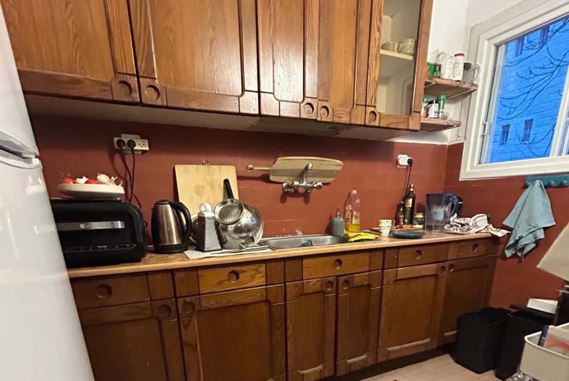 Cozy kitchen with wooden cabinets, cluttered counter, toaster, kettle, sink, and dish rack scattered items under a red-brown backsplash.
