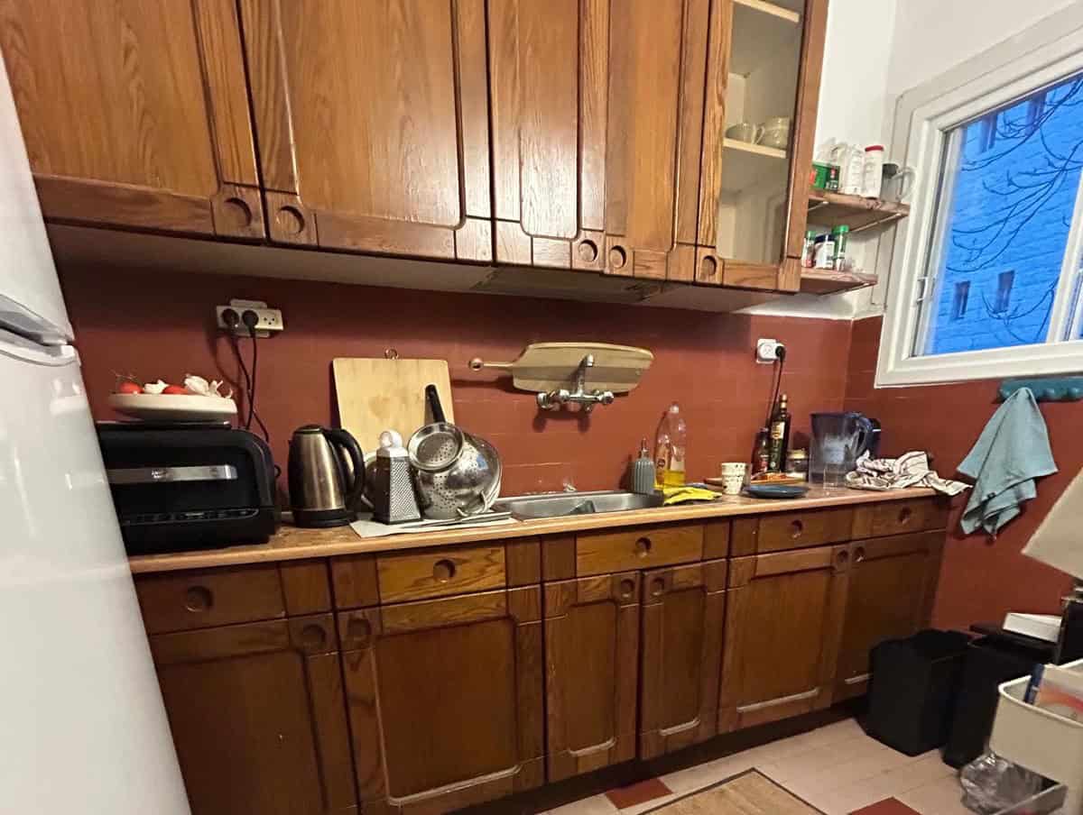 Cozy kitchen with wooden cabinets, cluttered counter, toaster, kettle, sink, and dish rack scattered items under a red-brown backsplash.