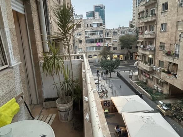 Balcony with potted plants overlooks a city street with outdoor cafe tables and white umbrellas, surrounded by beige apartment buildings.