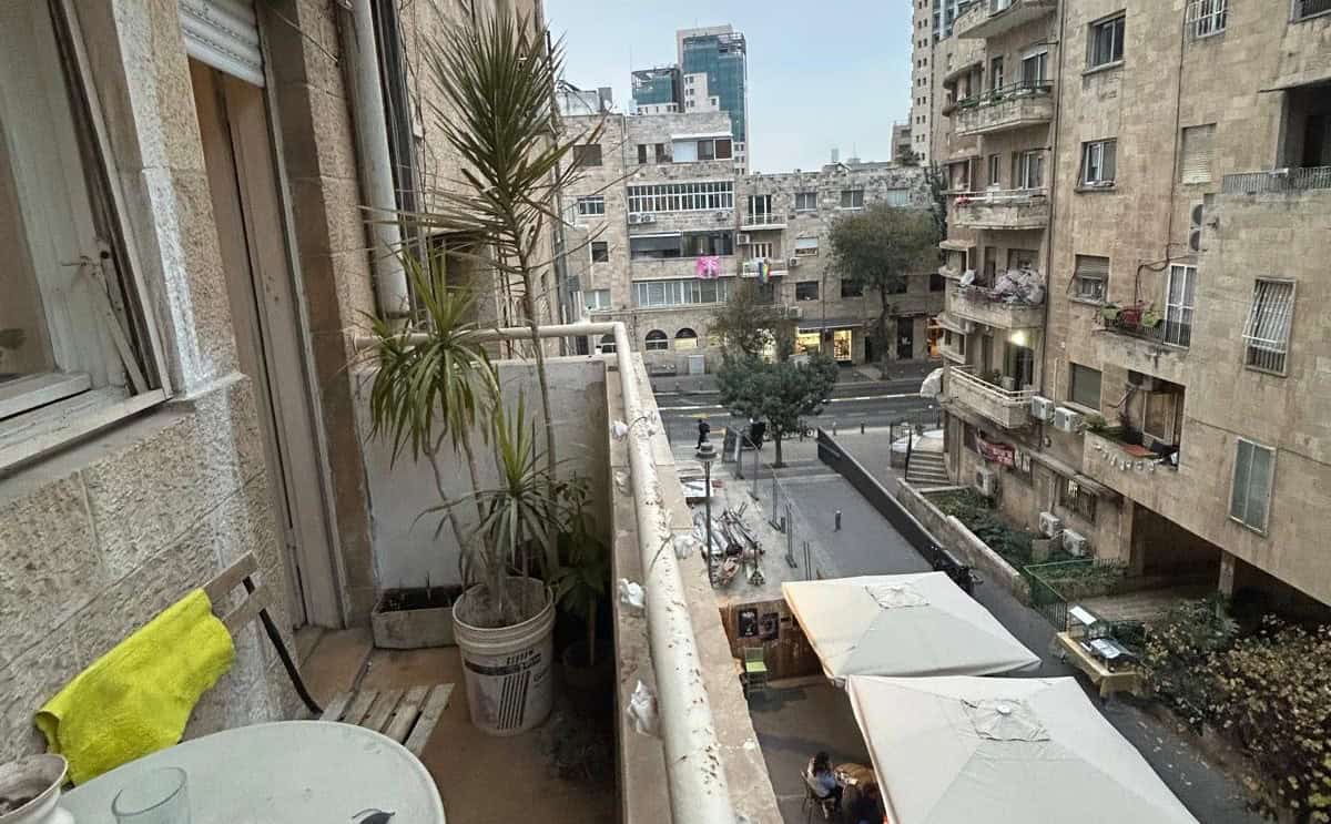Balcony with potted plants overlooks a city street with outdoor cafe tables and white umbrellas, surrounded by beige apartment buildings.