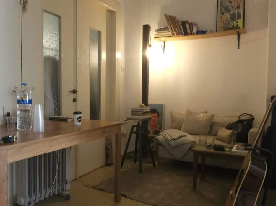 Cozy living space with a wooden table, water bottle, mug, and a couch cluttered with cushions and a stack of books nearby.