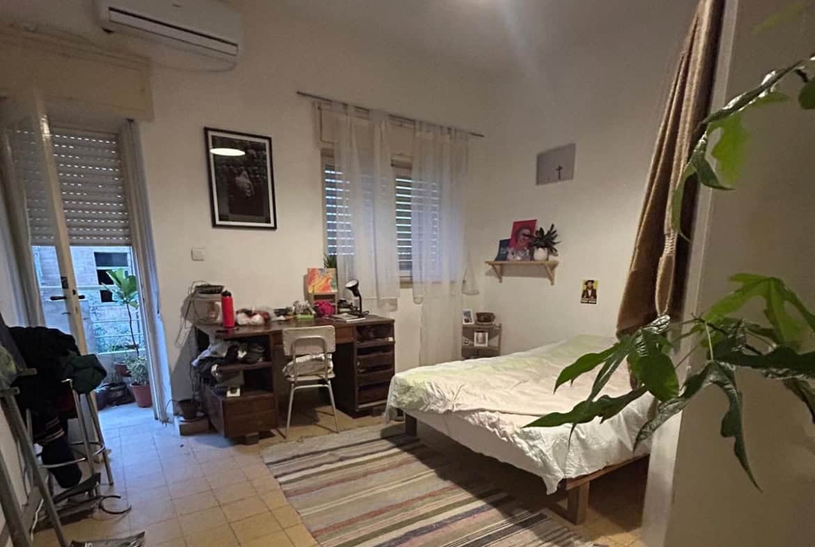 Bedroom with a single bed on the right, white bedding, a wooden desk with clutter and a metal chair, and a striped rug; a door to a balcony and a window let in light, with plants nearby.