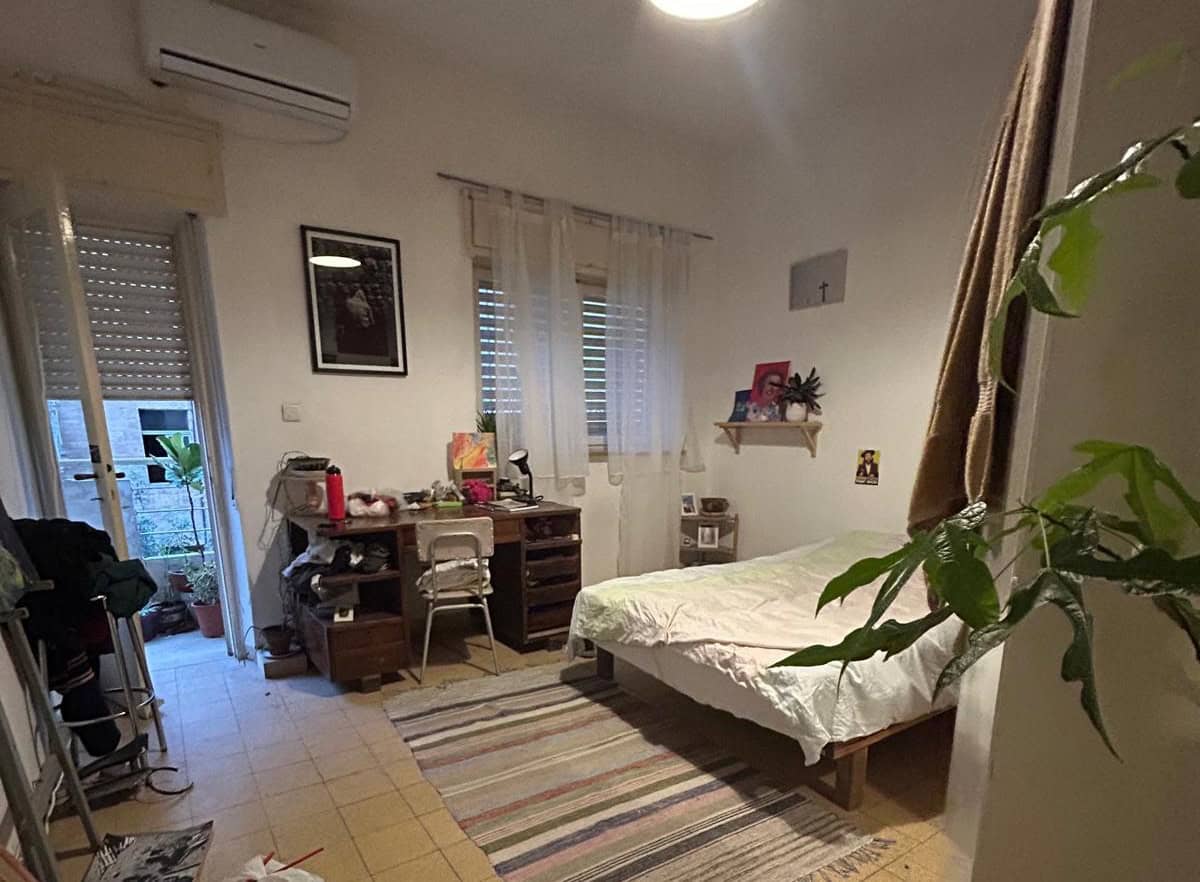 Bedroom with a single bed on the right, white bedding, a wooden desk with clutter and a metal chair, and a striped rug; a door to a balcony and a window let in light, with plants nearby.