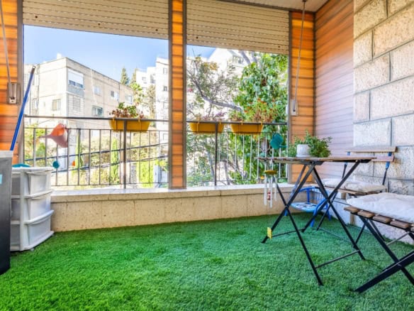 Balcony with green artificial grass, a folding table and bench, potted plants, and a railing overlooking nearby buildings.