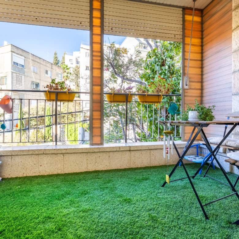 Balcony with green artificial grass, a folding table and bench, potted plants, and a railing overlooking nearby buildings.