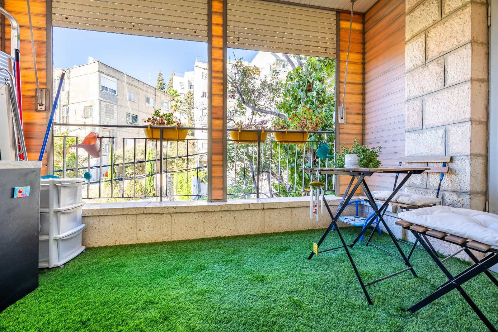 Balcony with green artificial grass, a folding table and bench, potted plants, and a railing overlooking nearby buildings.