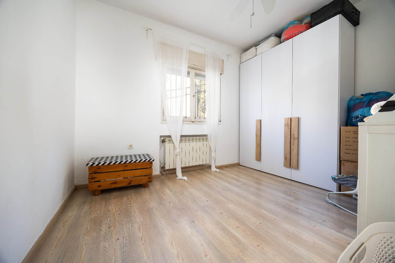 Bright, sparsely furnished bedroom with a large white wardrobe, a window with sheer curtains, and a small wooden bench by the radiator.
