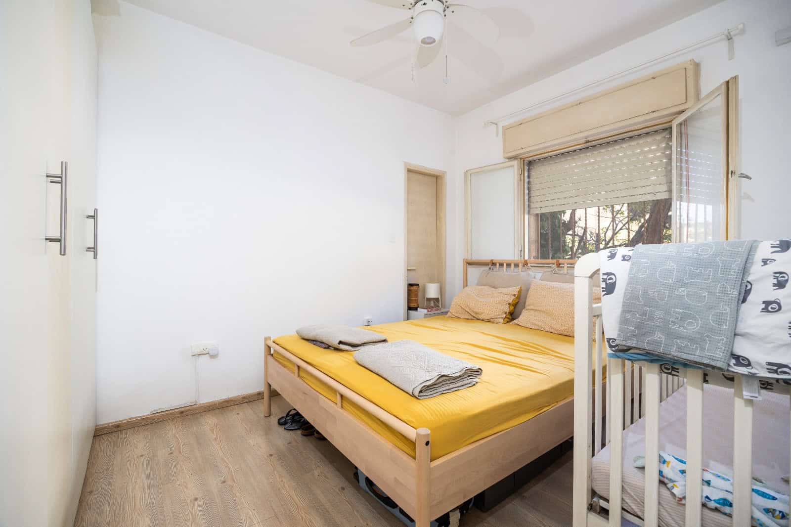 A small bedroom with a wooden double bed, yellow bedding, and folded blankets at the foot, next to a white bunk bed by the window.
