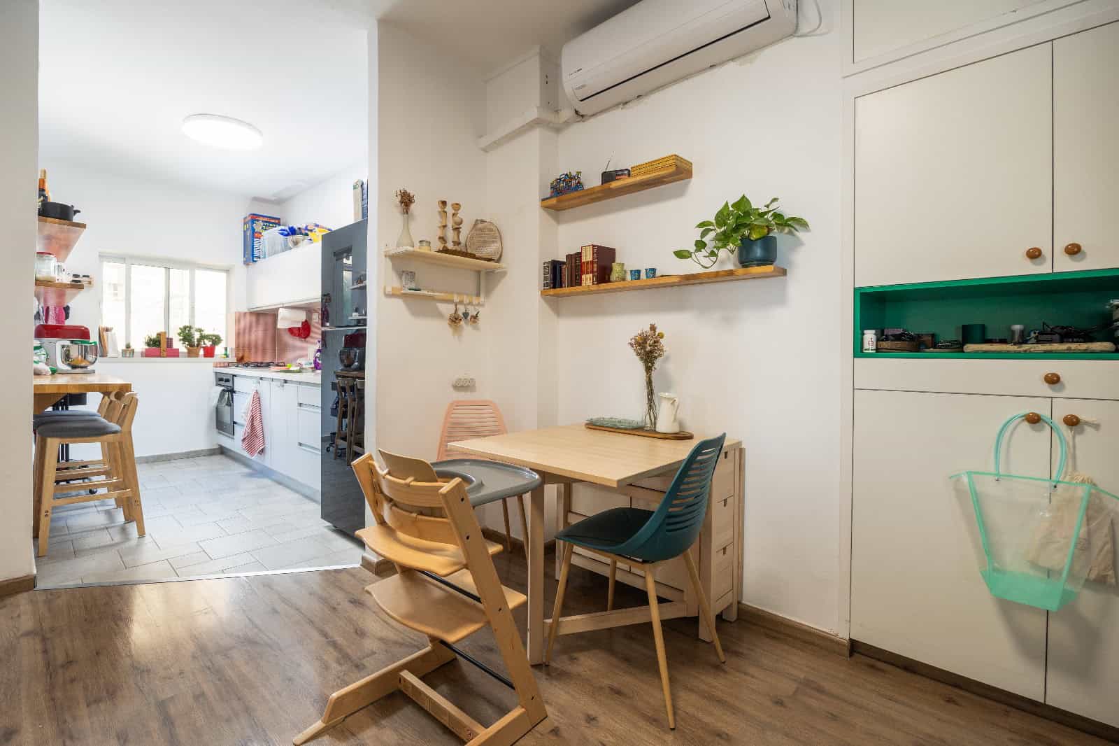 Open-plan kitchen and dining area with a wooden table and mixed chairs, white cabinets, and decorative shelves on light walls.